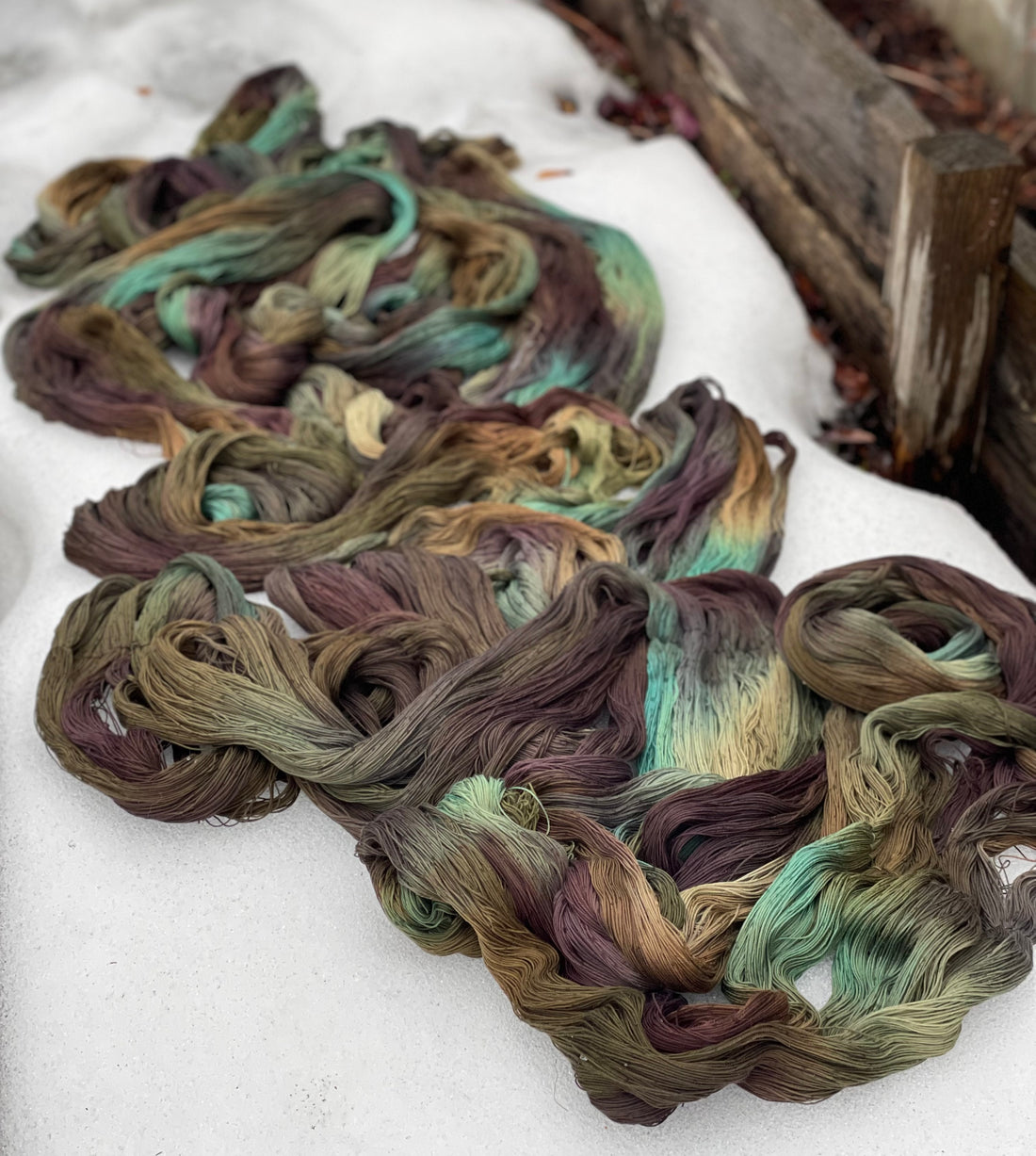 Image of a Cotton warp, laid out on the snow. Warp is dyed in shades of sage/browns/bronze/gold with pops of Seafoam and Ecru. 